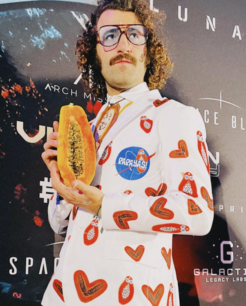 Alejandro Glatt in a white papaya-print suit, holding a halved papaya at a space-mission press wall